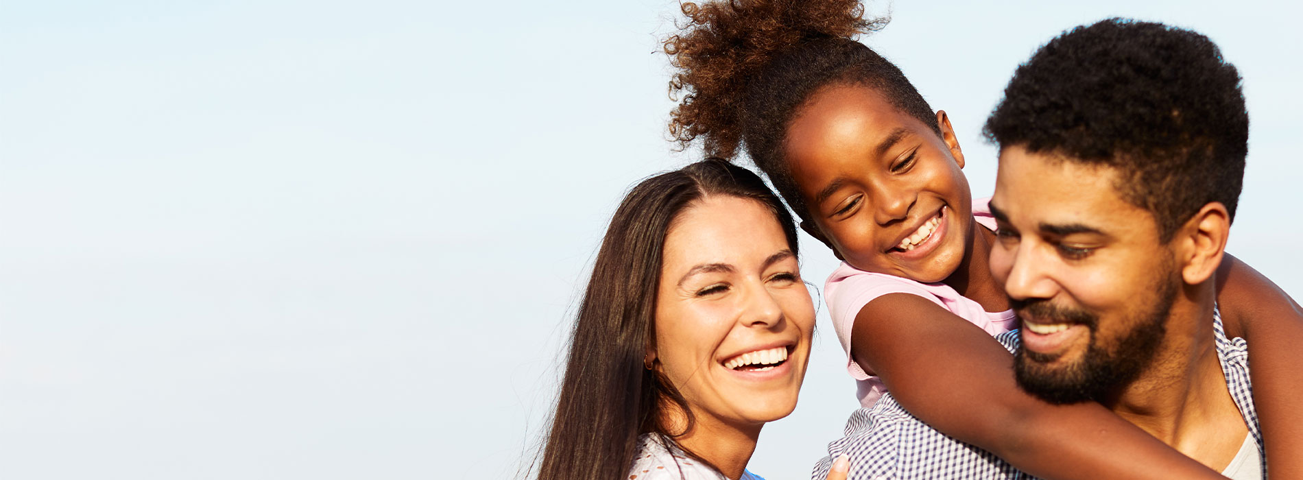 The image depicts a family of three, with two adults and a child, embracing each other outdoors during the day.